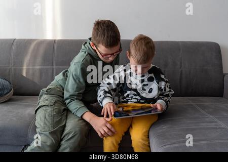 Two brothers sitting on grey sofa playing together on tablet with older boy helping younger sibling at home. Concept of siblings bonding, digital lear Stock Photo