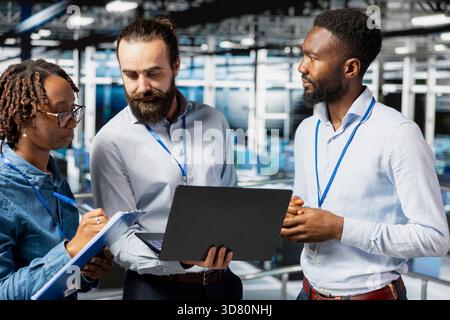 Data center team leader and workers reviewing performance dashboards of AI equipment rigs. Server room engineers overseeing machine learning algorithms on laptop, elevated workstation. Stock Photo