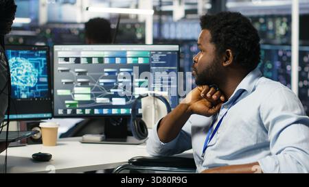 Data center engineer in wheelchair and colleague oversee automated tasks using node tree programming tool. Inclusive server farm team adjusts system settings based on AI analytics, camera B Stock Photo