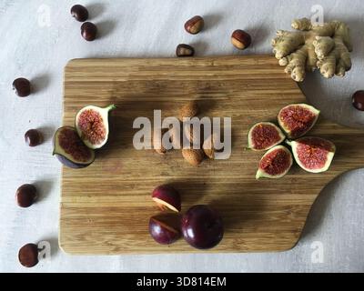 Top-down view of a wooden cutting board displaying almonds, plums, figs, ginger pieces and chestnuts. The ingredients create a natural autumn vibe. Stock Photo