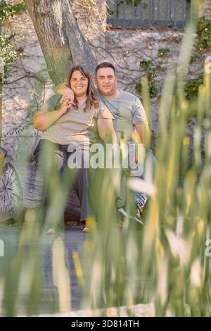 Young Christian couple sitting on a tree log outdoors, surrounded by greenery. Overweight man and woman share a relaxed and intimate moment in natural Stock Photo