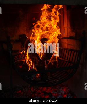 Vertical shot of a powerful, vibrant fire burning in a dark indoor fireplace or grill, with logs resting on a metal grate above glowing red embers. Pe Stock Photo