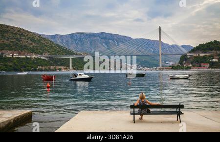 Young blond woman sitting on a bench and looking at the Tudjman Bridge in Dubrovnik, Croatia Stock Photo