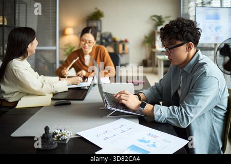 Young Asian businessmen and businesswomen working together in modern office, young Asian man using laptop at desk while two young Asian women discussing documents and charts in background Stock Photo