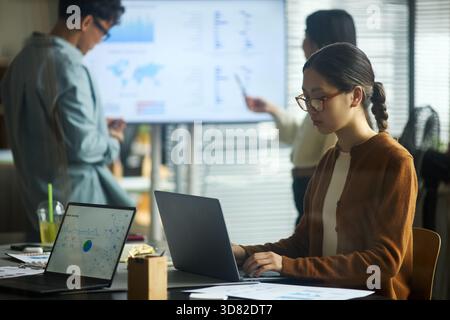 Young Asian businessmen and businesswomen working in modern office, young Asian woman using laptop at desk while two young Asian colleagues discussing data on digital screen in background Stock Photo