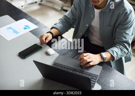 Young Asian man working on laptop at office desk, using wireless earphones and smartphone, analyzing business documents with charts, wearing smartwatch, focused on professional tasks Stock Photo