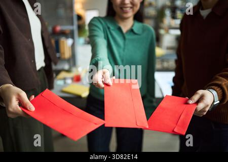 Young Asian businessmen and businesswomen holding red envelopes and standing together in office, smiling and extending hands forward, partial bodies visible, focus on gesture Stock Photo