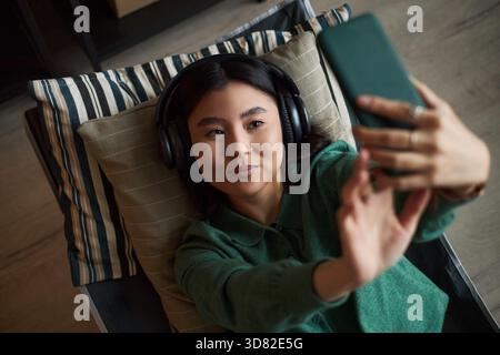 Portrait of young adult Asian woman lying on couch wearing headphones holding smartphone taking selfie smiling slightly relaxed expression modern office environment Stock Photo