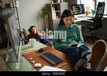 Young Asian woman sitting at desk using smartphone with legs on table, while young Asian man lying on floor in background wearing headphones in modern office workspace setting Stock Photo