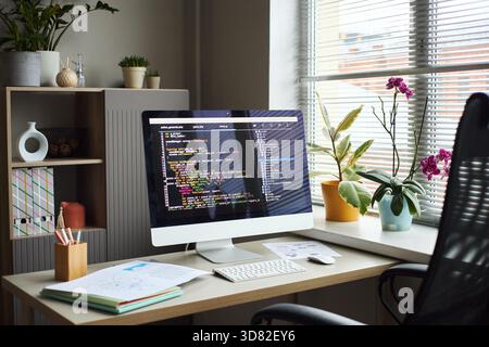 Young Asian man working at desk using desktop computer with programming code on screen, surrounded by office plants and documents, modern office environment visible through window blinds Stock Photo