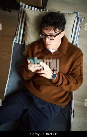 Young adult Asian man lying on office lounge chair using smartphone, wearing glasses and smartwatch, relaxing during work break, looking at device screen, casual business setting Stock Photo