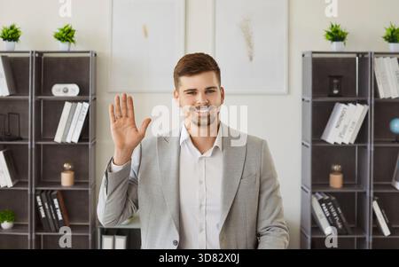 Young man looking at camera waving hand having video call standing on workplace in office. Stock Photo