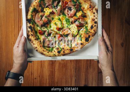 Two artisanal pizzas with assorted toppings served in open delivery boxes on a wooden table, viewed from above for commercial use. Stock Photo
