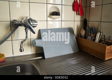 Plastic cutting board drying on a sink against a cream tile backsplash in a cluttered kitchen. Everyday real-life home scene. Concept of domestic, hom Stock Photo