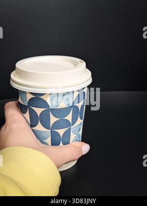 Close-up of a hand holding a takeaway coffee cup with a geometric pattern on a dark background Stock Photo