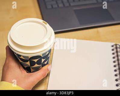 Close-up of a hand holding a coffee cup next to an open notebook and a laptop on a wooden desk Stock Photo