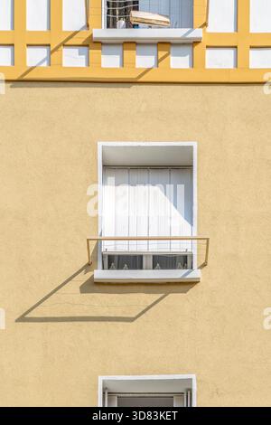 Detail of a yellow stucco residential facade with a closed window and strong shadow lines from the window railing and trim Stock Photo