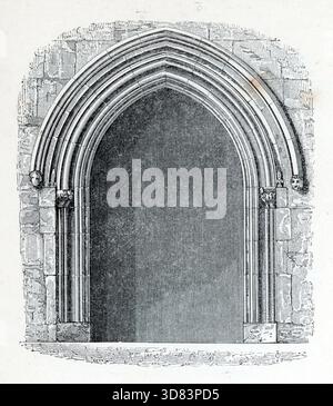 The monochrome engraving shows a grand, deeply recessed Gothic archway set into a stone block wall. The pointed portal features prominent multi-layered concentric moldings supported by capital columns, with the outer hoodmold terminating on either side in small, carved human head stops. From one of a collection of woodcuts published in the 4th (enlarged) edition of A Glossary of Terms Used In Grecian, Roman, Italian and Gothic Architecture, which was published in 1845. In some cases the pages had some moderate foxing, which may be visible in my photographs. Stock Photo