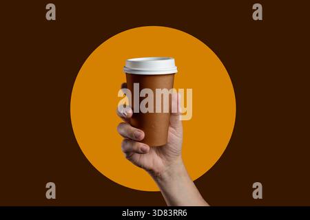 A man's hand holds a brown paper coffee cup against a modern graphic design with an orange circle, isolated on a brown background. Stock Photo