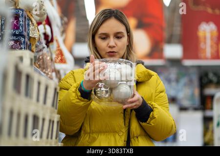 Woman holding glass ornament smiling in bright store aisle wearing yellow puffer jacket, inspecting sparkling bauble near decorated evergreen trees, f Stock Photo