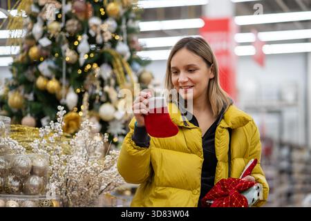 Caucasian woman examining red stocking, holding wrapped gift, golden decorated tree in background, checking texture and size, thoughtful smile, indoor Stock Photo