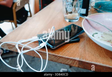 a smartphone with its white charging cable connected to a power bank rests on a wooden restaurant table, beside an empty glass and a bowl with food re Stock Photo