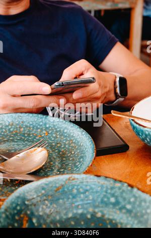 a man seated at an outdoor restaurant table uses his smartphone while it remains plugged into a black power bank, with empty blue plates and cutlery s Stock Photo