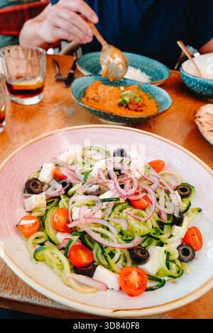 a bowl filled with spiralized cucumber mixed with cherry tomato halves, black olives, diced cheese and sliced red onion sits on a wooden table while a Stock Photo