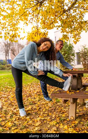 Mature couple warming up with leg stretches in a park covered with fallen yellow leaves Stock Photo