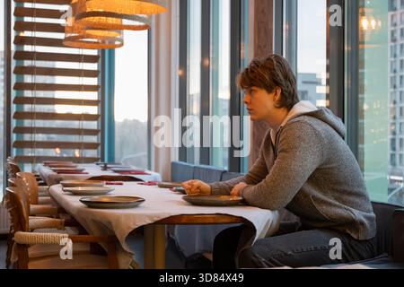 Young man wearing casual clothes sits alone at table near the window. The setting is cozy with empty tables around. Coffee Break at Cafe Stock Photo