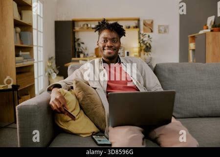 Portrait of Young Adult Black Man Shopping Online Using Laptop on Sofa Stock Photo