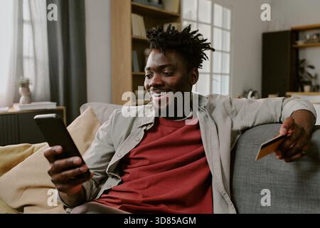 Young Black Man Sitting on Sofa Using Smartphone for Online Shopping Stock Photo