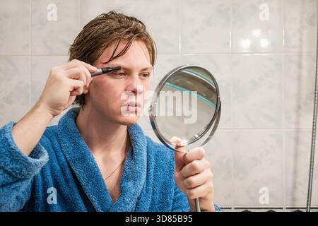 Young caucasian man in blue bathrobe using an electric trimmer to shape his eyebrows while looking into mirror in bathroom. Mens Home Self-Care Routin Stock Photo
