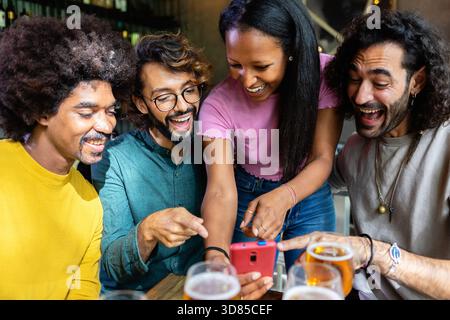 Happy young multiracial friends drinking beer and using smart phone at bar Stock Photo