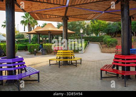 Bright round benches painted in rainbow colors under umbrella-shaped wooden canopies in a public park with greenery and walking paths. Stock Photo