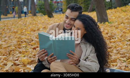 Hispanic couple in autumn park picnic reading book together communicate sitting in yellow leaves on blanket outdoors romantic date Arabian man with Stock Photo