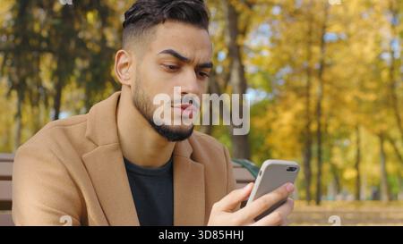 Portrait serious frown Hispanic man using mobile phone guy use app in autumn park reading bad news chat pensive negative emotion relaxing on bench in Stock Photo