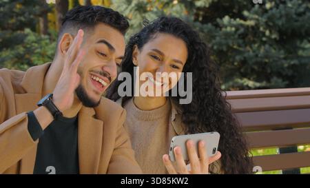Happy hispanic couple talking video call with mobile phone in autumn city park sitting on bench family smiling Arabian woman man online talk Stock Photo