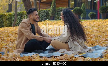 Happy Hispanic couple on date in autumn park talking laughing having fun sitting on blanket on yellow foliage Arabian man woman in love talk smile Stock Photo