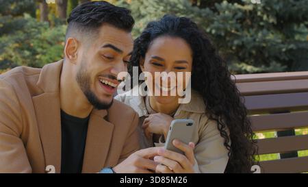 Hispanic happy couple guy with girl in autumn park on bench looking in phone cheerful family man woman discussing shopping online booking restaurant Stock Photo