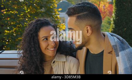 Cute Hispanic couple covered with plaid blanket date in autumn park on bench in city smiling cuddling talking love dating affectionate Caucasian woman Stock Photo