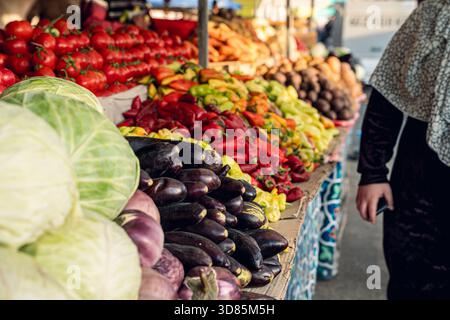 A colorful array of fresh vegetables at a bustling market, featuring tomatoes, peppers, and eggplants, with a shopper in the foreground. Stock Photo