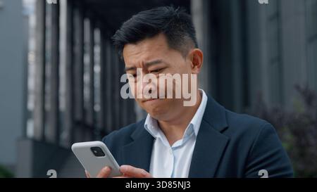 Middle-aged Asian businessman in city with phone sad Korean business man with mobile app failure upset worried looking in smartphone Chinese Japanese Stock Photo