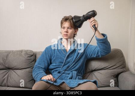 Young caucasian man in blue bathrobe using black hairdryer to dry his hair while sitting on sofa. Mens Home Self-Care Routine Stock Photo