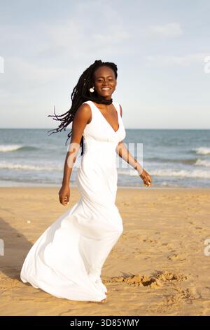 Young african american woman with dreadlocks in a flowing white dress,  barefoot and smiling as she walks confidently along a sunlit sandy beach  beside Stock Photo - Alamy