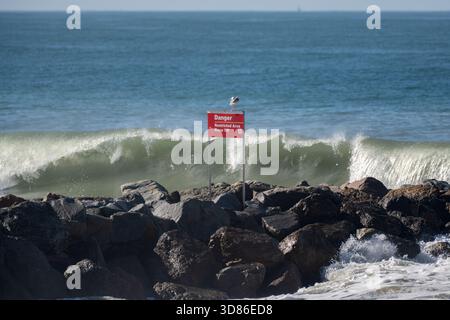 Danger sign warns as large waves break over the rock jetty in Pierpont Bay as a seafull looks on. Stock Photo