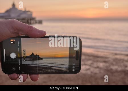 Hand holding a mobile phone taking a photo of a Pier at sunrise Stock Photo