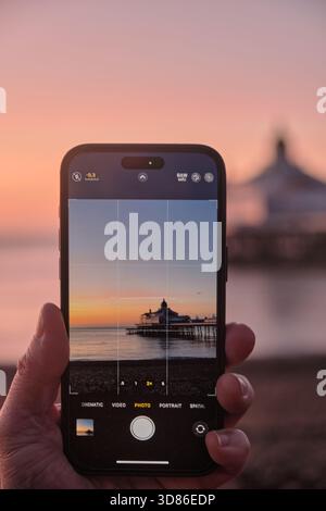 Hand holding a mobile phone taking a photo of a Pier at sunrise Stock Photo