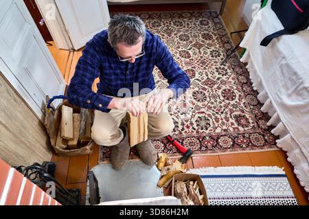 Shaving kindling from firewood log, homeowner prepares small wood strips for lighting brick stove in country house with woodburning heater. Stock Photo