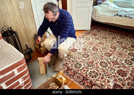Kindling preparation for brick heating unit, middle aged man seated on low stool uses small hatchet to chip fine wood slivers before igniting firewood Stock Photo